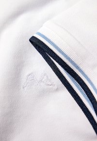 Weißes Polohemd aus Stoff mit strukturierter Oberfläche. Verfügt über einen Kragen mit navy- und hellblauem gestreiften Besatz sowie einem gestickten Logodetail.