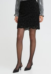 Zwarte kant rok met gegolfde zoom, gecombineerd met een metallic zilveren blouse. Het model draagt doorschijnende zwarte panty’s en zwarte hoge hakken.