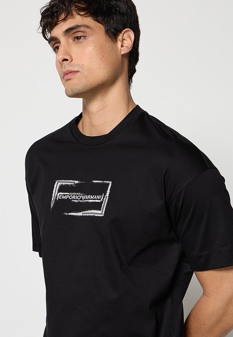 Hombre con camiseta negra de Emporio Armani con logo rectangular blanco, mirando ligeramente hacia la izquierda contra un fondo claro y liso.