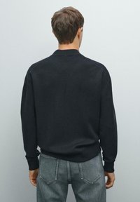 Svart stickad tröja med långa ärmar och ribbad krage. Bärs tillsammans med ljusgrå jeans, visad från baksidan mot en neutral bakgrund.