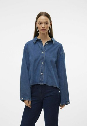 Blå denimskjorta med krage, knäppt fram och vida ärmar. Avslutad med silverknappar och en kortad fåll, ihop med mörka denimbyxor.