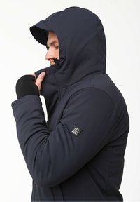 Chaqueta aislante de color navy con capucha, que presenta un cierre de cremallera y agujeros para los pulgares, fabricada con un tejido suave y con un parche de logo en la manga izquierda.
