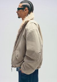 Khaki bombázó dzseki, prémes gallérral, oversized fazonnal és gumírozott mandzsettákkal. Alatta egy kockás inggel párosítva, oldalnézetből.