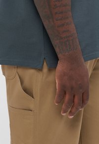Tume sinine lühikeste varrukatega särk, millel on jagatud serv, koos tumeroosade pükstega, millel on küljetasku ja sile tekstuur. Käsi on nähtav, millel on tätoveeringu detailid.