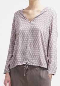Blouse légère à manches longues en rose pâle avec un motif de feuilles ; elle présente un col en V, un ourlet à cordon et une texture de tissu douce.