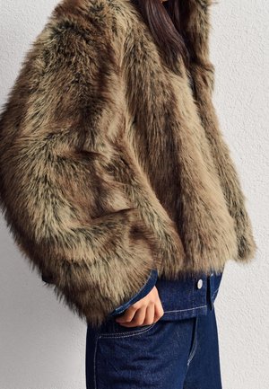 Personne portant une veste en fausse fourrure marron sur un pantalon en denim bleu foncé, debout contre un mur texturé clair.