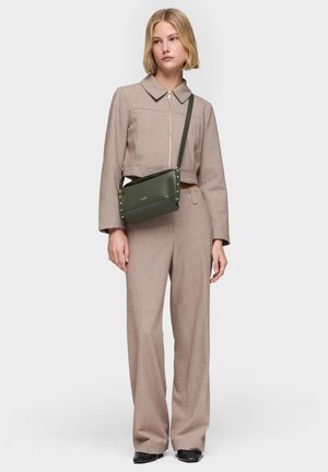 Mujer con chaqueta beige con cremallera y pantalones anchos a juego, llevando un bolso de hombro verde oliva con detalles dorados.