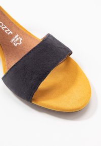 Avoin kärki sandaali, jossa on tummansininen samettinen päällinen ja keltainen samettinen pohja. Pohja on ruskeaa nahkaa, jossa on sileä rakenne. Minimalistinen muotoilu.