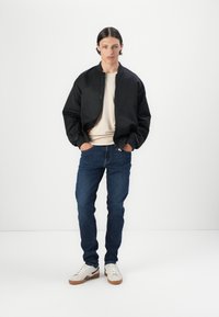 Chaqueta bomber negra, camisa beige debajo, jeans azules y zapatillas blancas con suelas marrones. Tejido suave, ajuste casual, sin patrones ni adornos.