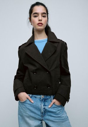 Mladá žena s tmavými vlasmi má na sebe čierny dvojradový kabát cez modrú košeľu a svetlomodré rifle, ruky vreckách, neutrálne pozadie.