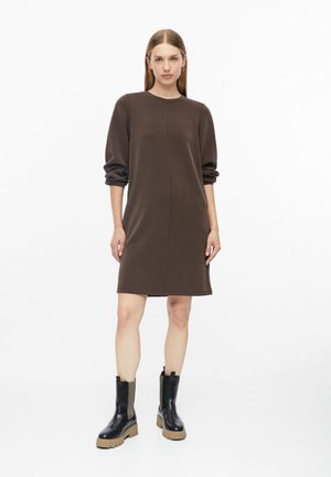 Femme debout portant une robe marron à manches longues et des bottines noires avec des semelles épaisses beige sur fond blanc.