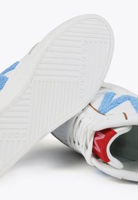 White sneaker featuring a blue textured detail, a red accent, and a rubber sole with a ridged pattern. Laces and perforated sections are visible.