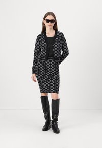 Cardigan à motifs noir et blanc sur une robe noire ajustée, associé à des bottes noires jusqu'aux genoux et des lunettes de soleil. Présente des boutons et un ourlet rayé.