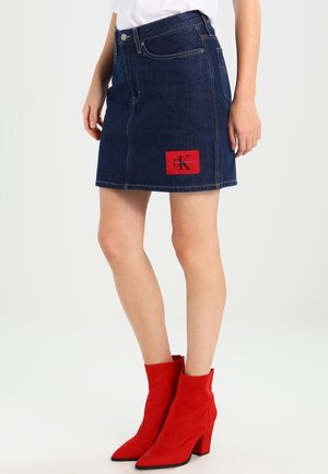 Vrouw draagt een donkerblauwe denim minirok met een rood logo-patch en rode suède enkellaarsjes met hoge hakken, staande tegen een effen achtergrond.