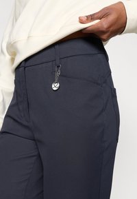 Tumed sinised püksid, mis on valmistatud siledast, venivast kangast. Pükstel on küljetaskud ja dekoratiivne hõbedane ripats, mis on kinnitatud vöökohtadele.