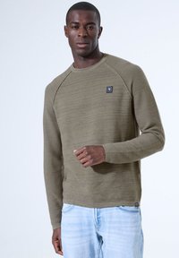 Olivenfarbene Langarm-Strickjacke mit strukturierten horizontalen Streifen, Rundhalsausschnitt und einem kleinen quadratischen Logo auf der Brust. Kombiniert mit hellblauen Jeans.