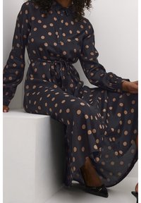 Lång svart klänning med stora beige prickar, knappframsida, krage och knytmidja. Tillverkad av ett lätt, flödande tyg.