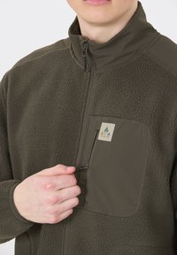 Olivgrön fleecjacka med en zippered bröstficka, strukturerad yttermaterial och slät krage. Har varumärkesetikett på fickan.