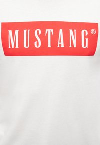 Balta medvilninė marškinėliai su ryškiu raudonu stačiakampiu logotipu, kurioje parašyta "MUSTANG" baltais rašmenimis. Logotipe yra registruotas prekių ženklas.