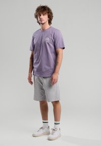 Light purple cotton t-shirt with a graphic print, paired with light gray shorts. Worn with white sneakers and striped socks, standing on a neutral background.