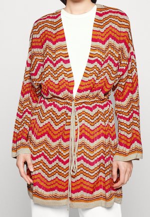 Cardigan en maille à manches longues avec des motifs en zigzag rose, orange et beige, noué à la taille, porté par-dessus un top blanc.
