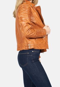 Giacca da moto in pelle marrone con spalle texture, zip asimmetrica e fibbie laterali, abbinata a jeans in denim blu scuro.