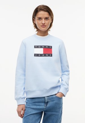 Ung person iført en lyseblå sweatshirt med et rødt, hvidt og navy Tommy Jeans-logo, kombineret med blå denimjeans.