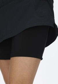 Svarta atletiska shorts gjorda av elastiskt material, med en slät yta och en åtsittande design, synliga under ett mörkt ytterplagg.