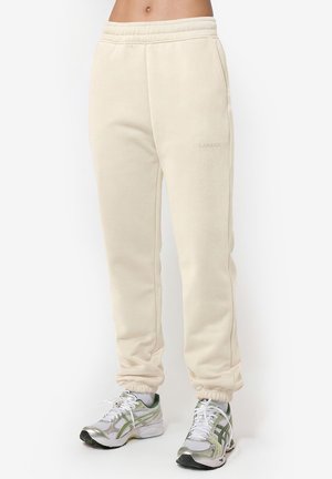 Pantaloni de trening bej din fleece, cu talie și mansete elastice, purtați cu pantofi de alergare albi și verzi pe o persoană în picioare.