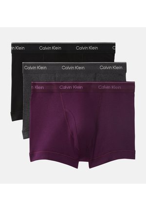 Ung mand står barfodet i mørkegrå Calvin Klein boxershorts med den ene hånd bag nakken mod en ensfarvet grå baggrund.