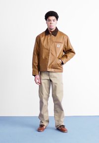 Giacca marrone, lucida e impermeabile con un colletto marrone, tasche frontali e un patch con logo, abbinata a pantaloni beige chiaro e scarpe marroni.