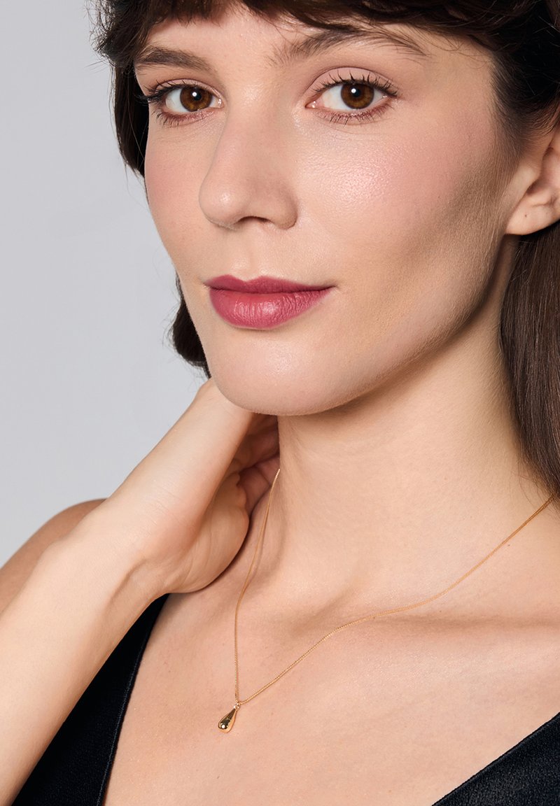 Złoty naszyjnik z zawieszką w kształcie kropli, cienki łańcuszek, noszony na nagiej szyi, na czarnej bluzce. Gładka tekstura i błyszcząca powierzchnia podkreślona.