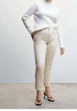 Donna che indossa un maglione bianco a coste, pantaloni beige a vita alta e sandali neri con tacco e cinturini, in piedi davanti a uno sfondo uniforme.