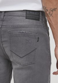 Jeans de mezclilla grises con una etiqueta de parche que lleva el texto "11 PROJECT". El bolsillo trasero presenta un sutil desgaste y tiene adherida una pequeña etiqueta de la marca.