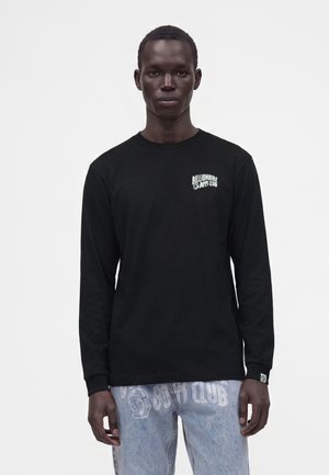 Uomo che indossa una maglia nera a maniche lunghe con il testo "Billionaire Boys Club" e jeans azzurri con loghi coordinati sulla parte anteriore.