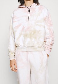 Hoodie tie-dye in rosa pastello e beige. Presenta una tasca frontale, cappuccio con cordino regolabile e polsini a costine. Tessuto morbido e texturizzato.