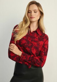 Czerwona koszula zapinana na guziki z wzorem akwarelowym, z długimi rękawami i kołnierzykiem, w zestawie z czarną skórzaną spódnicą. Gładka tekstura, dopasowany krój.