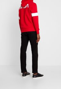 Červená mikina s kapucí s bílým logem NASA a pruhy na rukávech, v kombinaci s černými kalhotami a černými teniskami, pohled zezadu.