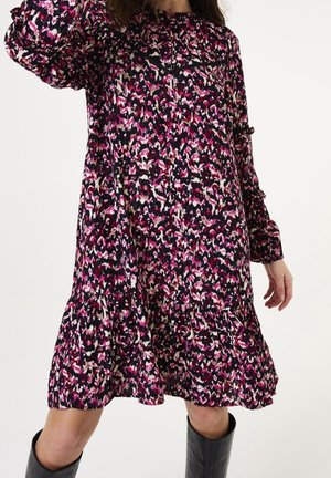 Nainen, jolla on päällään pitkähihainen, polvipituinen mekko, jossa on vaaleanpunaisia, mustia ja valkoisia abstrakteja kuvioita, yhdistettynä mustiin polven korkeisiin saappaisiin.