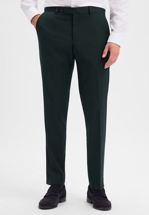 Man wearing dark green formal trousers with side pockets and black loafers standing against a plain background.