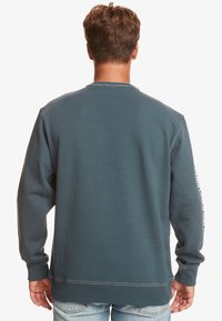 Felpa di cotone blu con collo a giro, polsini e fondo a costine; presenta testo bianco lungo la manica sinistra. Morbido al tatto.