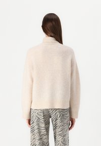 Jasno beżowy sweter z wysokim dekoltem, miękką fakturą, luźnym krojem i prążkowanym dołem, w zestawieniu z szerokimi spodniami w szaro-czarne paski przypominające tygrysa.