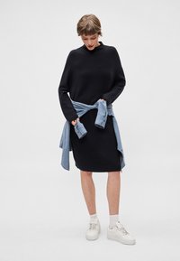 Černé pletené šaty s vysokým výstřihem a dlouhými rukávy, doplněné světle modrou košilí uvázanou v pase. Celkový vzhled dotvářejí bílé tenisky.