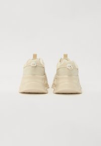 Beige sneakers with a smooth texture, featuring a cushioned sole, a pull tab at the heel, and decorative stitching along the sides.