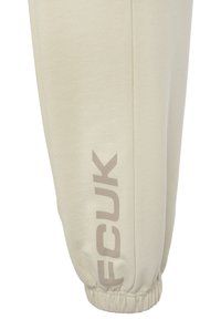 Beige joggerbroeken van zacht materiaal met een elastische boord. Voorzien van een groot "FCUK" logo in lichtbruin op het linkerbeen.