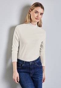 Långärmad, höghalsoverdel i mjukt krämfärgat tyg, kombinerad med mörkblå denimjeans. Slät textur med figursydd design och uppvikta ärmar.