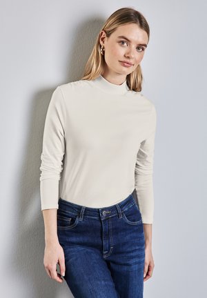 Camiseta de manga larga y cuello alto en suave tela crema, combinada con unos vaqueros de mezclilla azul oscuro. Textura suave con un diseño ajustado y mangas enrolladas.