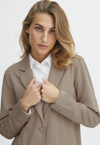 Beige blézer sima textúrával, kiöltött gallérral, két elülső gombbal és alatta egy fehér inggel. Kezek finoman összekulcsolva a gallérnál.