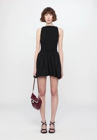 Black sleeveless dress with a smocked bodice and flared skirt. Model carries a burgundy bag and wears strappy black sandals.