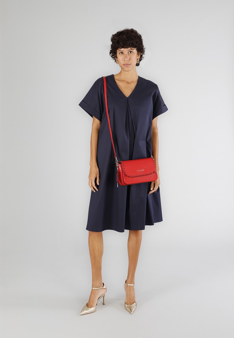 Abito blu navy con scollo a V e maniche corte; tessuto liscio. Borsa a tracolla rossa con dettagli intrecciati. Scarpe con tacco dorato.
